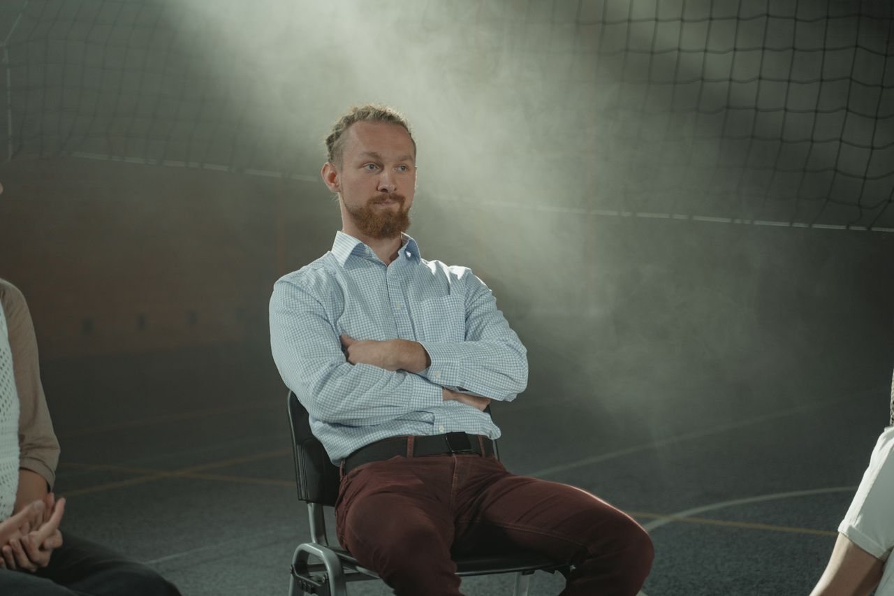 A man sits pensively during a group therapy session, illuminated by soft, filtered light indoors.
